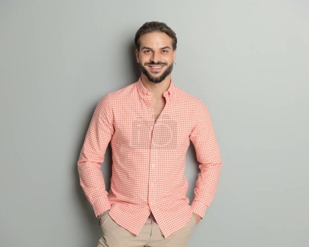 Foto de Feliz hombre casual en camisa a cuadros tomados de la mano en los bolsillos y sonriendo mientras posando delante de fondo gris - Imagen libre de derechos