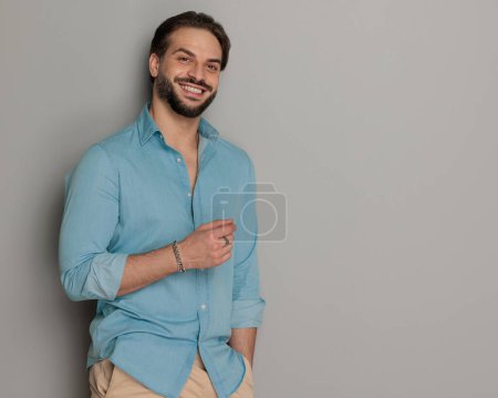 Téléchargez les photos : Attrayant jeune homme avec barbe souriante et tenant la main dans la poche devant fond gris - en image libre de droit
