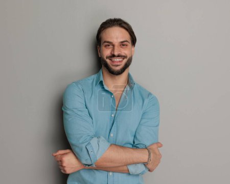 Photo for Happy young man in denim shirt smiling and crossing arms while posing in front of grey background - Royalty Free Image