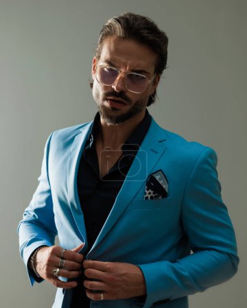 Téléchargez les photos : Portrait de cool homme élégant avec des lunettes posant tout en boutonnant costume et être confiant devant fond gris - en image libre de droit