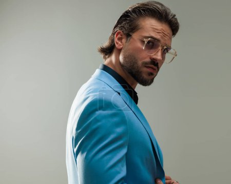 Foto de Vista lateral del elegante hombre de negocios con gafas ajustando el traje y mirando hacia adelante en frente de fondo gris - Imagen libre de derechos