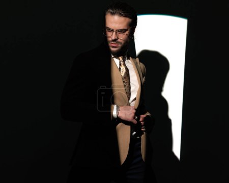 Photo for Elegant high class man with glasses looking away while closing suit in front of window shadow on grey background - Royalty Free Image