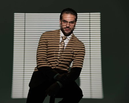 Téléchargez les photos : Homme d'affaires élégant sexy avec des lunettes tenant les coudes sur les genoux, regardant vers l'avant et assis devant l'ombre de la fenêtre sur fond gris - en image libre de droit