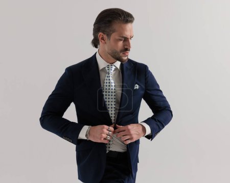 Foto de Retrato de elegante hombre de negocios de alta clase abotonando traje azul marino y mirando hacia un lado en frente de fondo gris - Imagen libre de derechos