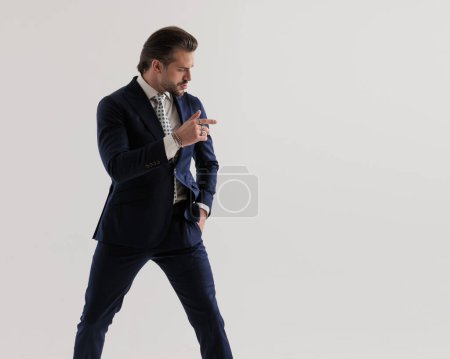 Foto de Fresco hombre de negocios elegante en traje azul marino mirando y apuntando de un lado a otro mientras posando con una mano en el bolsillo sobre fondo gris - Imagen libre de derechos