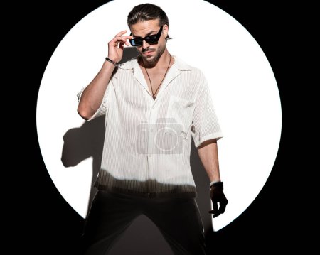 Téléchargez les photos : Cool casual homme regardant vers le bas et ajustant les lunettes de soleil d'une manière confiante devant projecteur rond sur fond blanc en studio - en image libre de droit