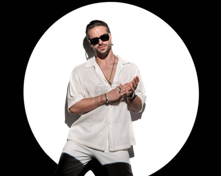 Téléchargez les photos : Portrait d'un homme barbu confiant avec des lunettes de soleil regardant vers l'avant et frottant les paumes devant un projecteur rond sur fond blanc - en image libre de droit