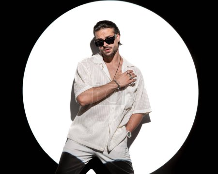 Foto de Atractivo hombre casual con gafas de sol, sosteniendo el brazo en pose de moda y posando con una mano en el bolsillo delante de proyector redondo sobre fondo blanco - Imagen libre de derechos