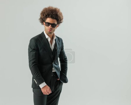 Téléchargez les photos : Homme d'affaires confiant en smoking noir avec chemise col ouvert tenant la main dans des poches devant fond gris - en image libre de droit