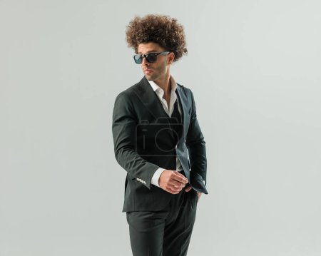 Téléchargez les photos : Sexy élégant homme d'affaires tenant la main dans la poche et regardant de côté tout en posant d'une manière confiante sur fond gris - en image libre de droit