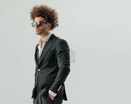 Foto de Sexy hombre de negocios afro con el pelo largo rizado mirando hacia otro lado y posando con las manos en los bolsillos delante de fondo gris - Imagen libre de derechos