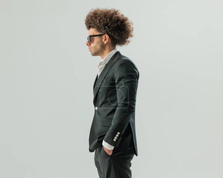 Téléchargez les photos : Vue latérale de l'homme élégant avec des cheveux afro et des lunettes de soleil tenant la main dans les poches et regardant de côté sur fond gris - en image libre de droit