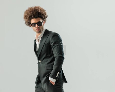 Téléchargez les photos : Portrait d'un jeune homme élégant avec des lunettes portant une chemise à col ouvert avec smoking noir et tenant la main dans des poches sur fond gris - en image libre de droit
