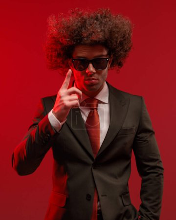 Foto de Sexy joven en traje con corbata roja sosteniendo la mano en el bolsillo y apuntando con el dedo hacia arriba en frente de fondo gris - Imagen libre de derechos