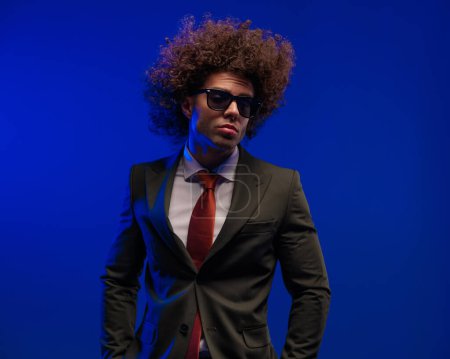 Téléchargez les photos : Cool afro homme d'affaires en costume noir avec des lunettes de soleil tenant la main dans les poches et posant devant fond bleu - en image libre de droit
