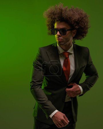 Téléchargez les photos : Portrait de sexy homme élégant avec des lunettes de soleil regardant de côté tout en déboutonnant costume en face de fond vert - en image libre de droit