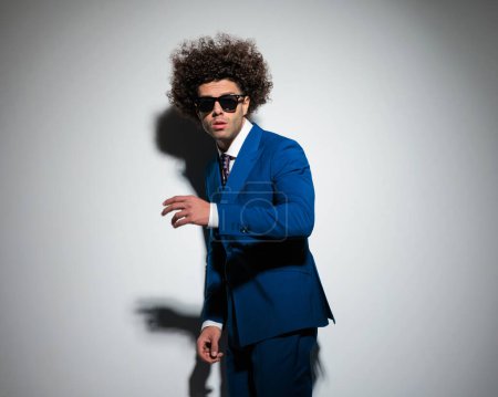Photo for Cool fashion businessman with long curly hair and sunglasses posing with arms in a fashion pose in front of grey background - Royalty Free Image