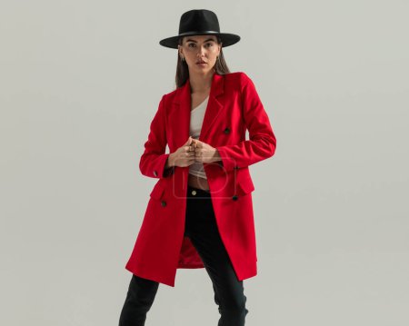 Photo for Cool fashion woman with hat closing and adjusting red coat while posing in a confident way in front of grey background - Royalty Free Image