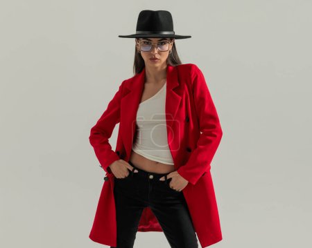 Téléchargez les photos : Coussin femme confiant avec chapeau et manteau rouge tenant les mains dans les poches et étant cool devant fond gris - en image libre de droit