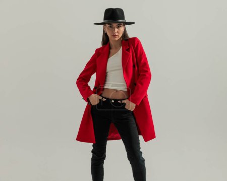Foto de Sexy chica con el pelo largo y sombrero con abrigo rojo y la celebración de las manos en los bolsillos delante de fondo gris - Imagen libre de derechos