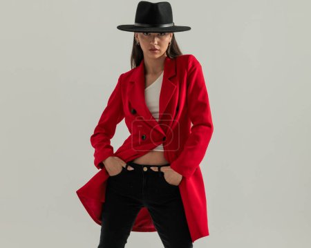 Téléchargez les photos : Belle femme décontractée avec chapeau en manteau rouge tenant les mains dans les poches et posant devant fond gris - en image libre de droit
