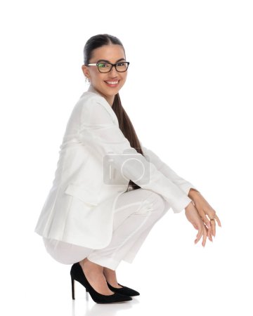 Téléchargez les photos : Vue de côté image de fille élégante heureuse avec des lunettes accroupi avec des coudes sur les genoux en face de fond blanc - en image libre de droit
