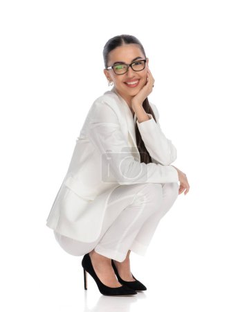 Téléchargez les photos : Attrayant jeune femme avec des lunettes accroupi et tenant menton dans les paumes tout en touchant les genoux avec les bras et en souriant sur fond blanc - en image libre de droit