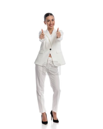 Téléchargez les photos : Image corporelle complète de heureuse jeune femme d'affaires souriant tout en faisant les pouces vers le haut geste devant fond blanc - en image libre de droit