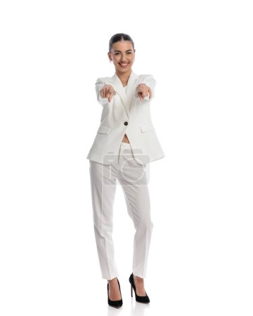 Téléchargez les photos : Excité élégant jeune femme en costume blanc souriant tout en pointant les doigts vers l'avant en face de fond blanc en studio - en image libre de droit