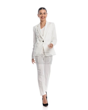 Téléchargez les photos : Image complète du corps de la femme d'affaires élégante en costume blanc souriant et marchant devant fond blanc - en image libre de droit