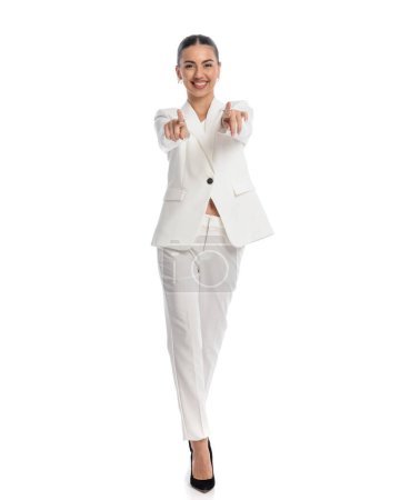 Téléchargez les photos : Heureuse femme élégante souriant tout en pointant les doigts vers l'avant et en marchant devant fond blanc - en image libre de droit