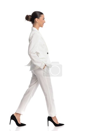 Téléchargez les photos : Sexy femme élégante en costume blanc avec des cheveux de chignon marchant avec les mains dans les poches et souriant devant fond blanc - en image libre de droit