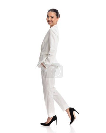 Foto de Vista lateral de elegante joven empresaria en traje blanco siendo feliz, sonriente y caminando con las manos en bolsillos delante de fondo blanco - Imagen libre de derechos