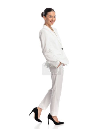 Téléchargez les photos : Vue latérale de jeune fille heureuse en costume blanc tenant la main dans les poches, souriant et marchant devant fond blanc - en image libre de droit
