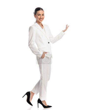 Foto de Orgullosa joven en traje blanco mostrando a un lado con una mano mientras sostiene la otra en el bolsillo y pisando fondo blanco - Imagen libre de derechos