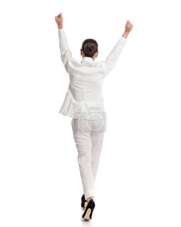 Téléchargez les photos : Vue arrière de la femme élégante en costume blanc tenant les bras au-dessus de la tête et acclamant tout en marchant devant fond blanc - en image libre de droit