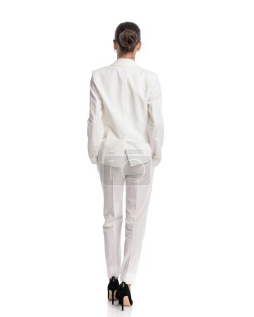 Foto de Vista trasera de la mujer sexy en traje blanco sosteniendo los brazos en los bolsillos y de pie delante del fondo blanco - Imagen libre de derechos
