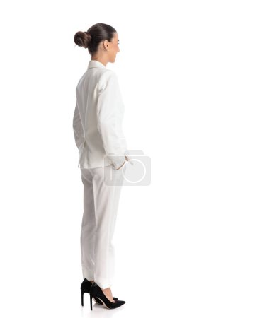 Téléchargez les photos : Vue latérale et arrière de la jeune femme élégante en costume blanc tenant la main dans les poches et regardant vers l'avant sur fond blanc - en image libre de droit