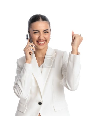 Photo for Happy elegant woman in white suit taling on the phone, laughing and cheering good news with fist up in front of white background - Royalty Free Image