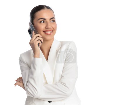 Foto de Hermosa mujer elegante en traje blanco tener una conversación telefónica, sonriendo y mirando hacia otro lado en frente de fondo blanco - Imagen libre de derechos