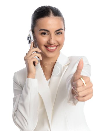 Photo for Portrait of attractive businesswoman talking on the phone and making thumbs up gesture while smiling in front of white background - Royalty Free Image