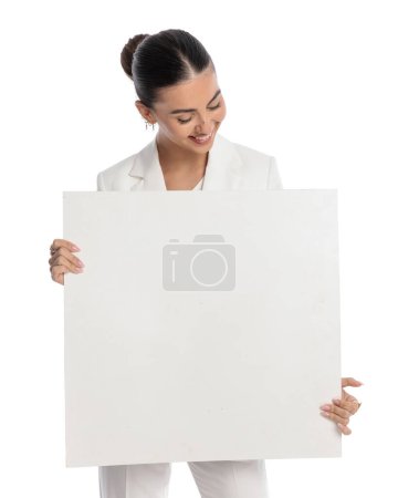 Téléchargez les photos : Portrait d'élégante femme de mode tenant panneau d'affichage, regardant vers le bas et souriant tout en présentant en face de fond blanc en studio - en image libre de droit
