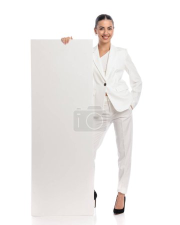 Photo for Confident woman in white suit showing and presenting empty board while standing with hand in pocket and smiling on white background - Royalty Free Image