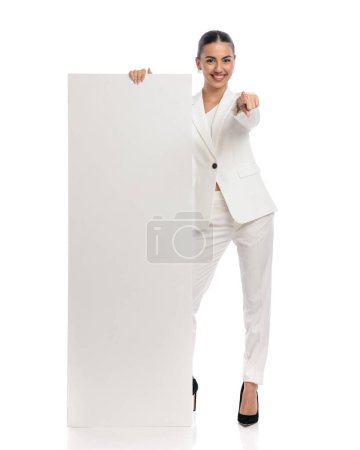 Photo for Beautiful businesswoman in white suit showing empty board, smiling and pointing finger forward in front of white background - Royalty Free Image