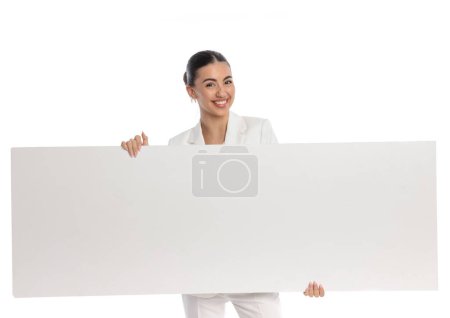 Téléchargez les photos : Séduisante femme d'affaires en costume blanc tenant et présentant tableau vide devant fond blanc - en image libre de droit