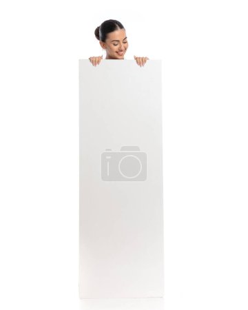 Téléchargez les photos : Femme heureuse regardant vers le bas et souriant tout en se cachant derrière la planche vide en face de fond blanc - en image libre de droit
