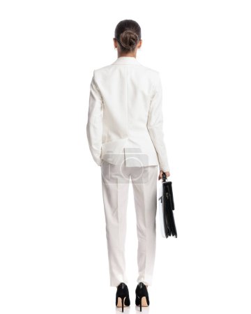Foto de Detrás de la vista de la joven y elegante mujer de negocios en traje blanco sosteniendo la maleta y de pie con la mano en el bolsillo delante del fondo blanco - Imagen libre de derechos
