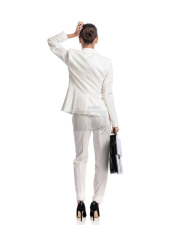 Téléchargez les photos : Vue arrière de l'élégante femme d'affaires en costume avec sac étant confus, la tête grattante et la pensée tout en se tenant debout sur fond blanc - en image libre de droit