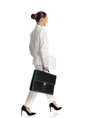 Téléchargez les photos : Attrayant jeune femme avec des cheveux de chignon en costume tenant le sac et marchant devant fond blanc - en image libre de droit