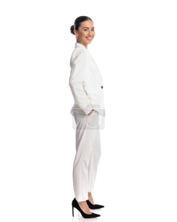 Foto de Vista lateral de feliz mujer de negocios elegante cogida de la mano en los bolsillos y sonriendo delante de fondo blanco - Imagen libre de derechos